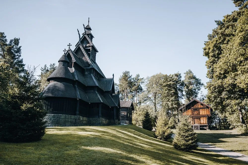 Die Stabkirche aus dunklem Holz mit Schindeldächern und mehreren Türmchen auf einer Wiese, dahinter Nadelbäume und ein braunes Holzhaus