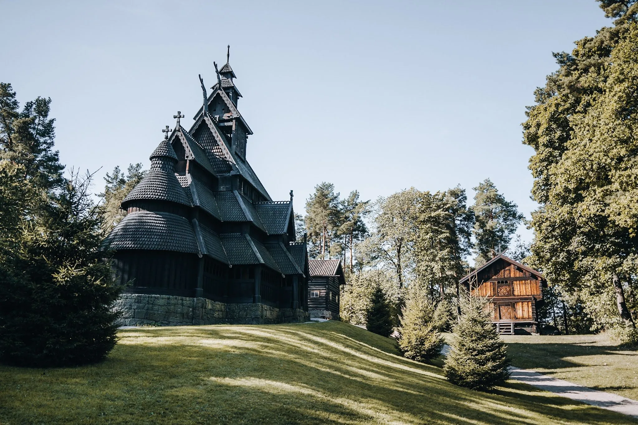 Die Stabkirche aus dunklem Holz mit Schindeldächern und mehreren Türmchen auf einer Wiese, dahinter Nadelbäume und ein braunes Holzhaus