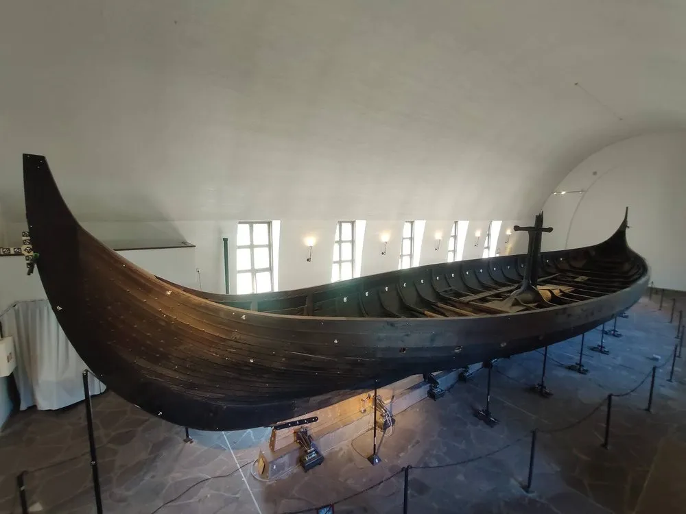 Ein Wikingerschiff aus dunklem Holz mit hoch geschwungenem Bug und Heck in einer Museumshalle, abgesperrt mit Stangen und Seilen