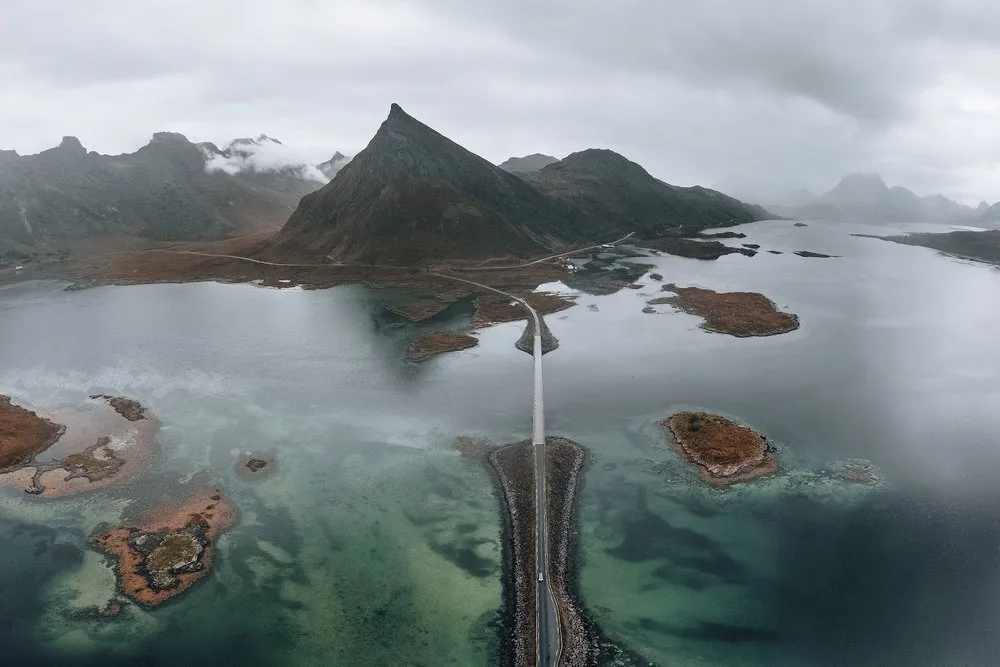 Luftaufnahme einer Strasse auf einem schmalen Damm über flaches, türkises Wasser mit kleinen Inseln, dahinter ein spitzer Berg im Nebel