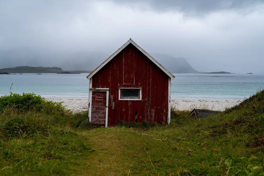 Die verwitterte rote Holzhütte steht zwischen Gras und Sandstrand am Rambergstranda, dahinter das Meer unter tiefen Wolken