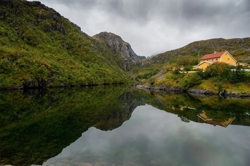 Ein gelbes Haus mit rotem Dach steht am Hang über spiegelglattem Wasser, links eine bewachsene Felswand