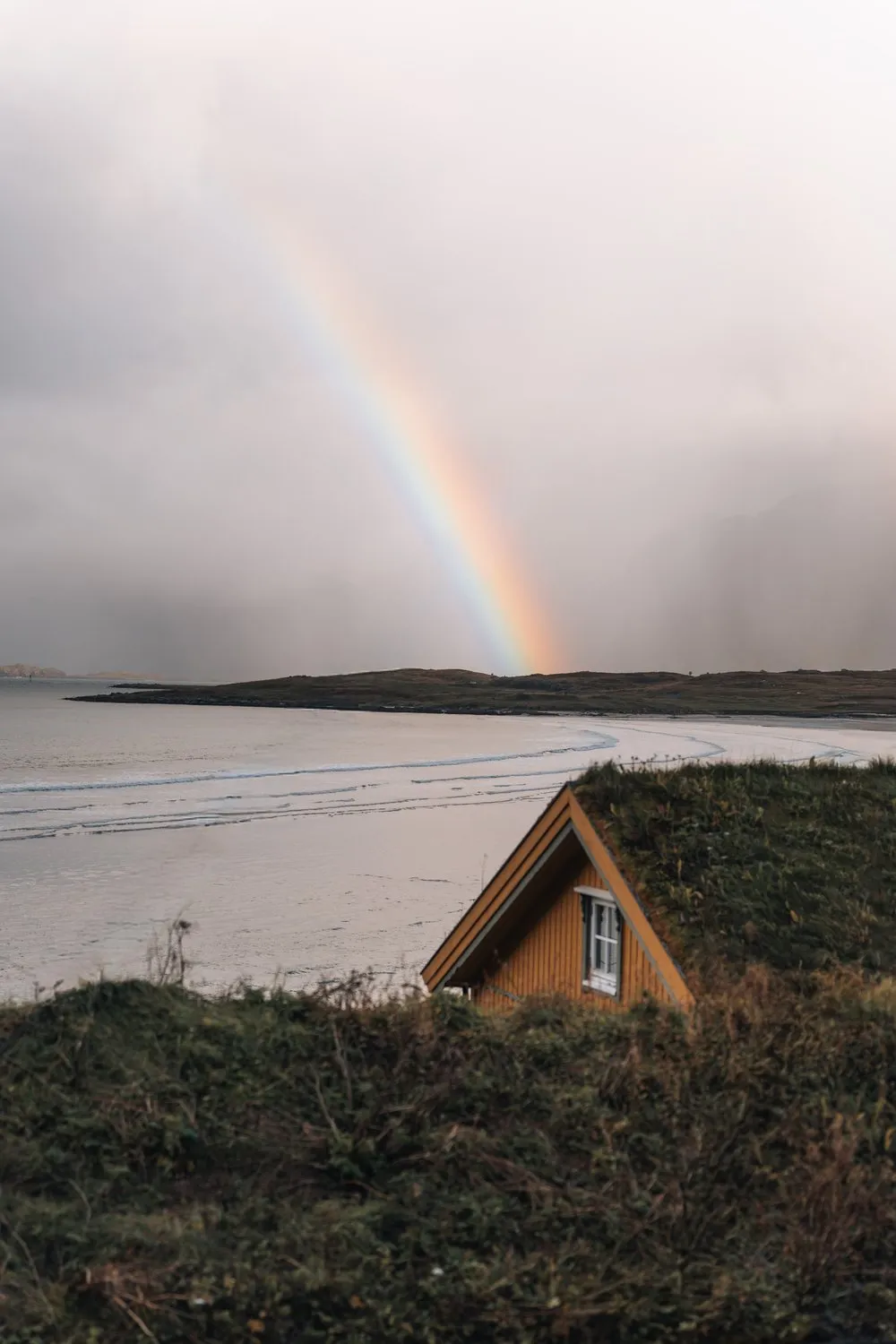 Ein Regenbogen über dem Strand von Fredvang, vorne ein kleines Haus mit Grasdach