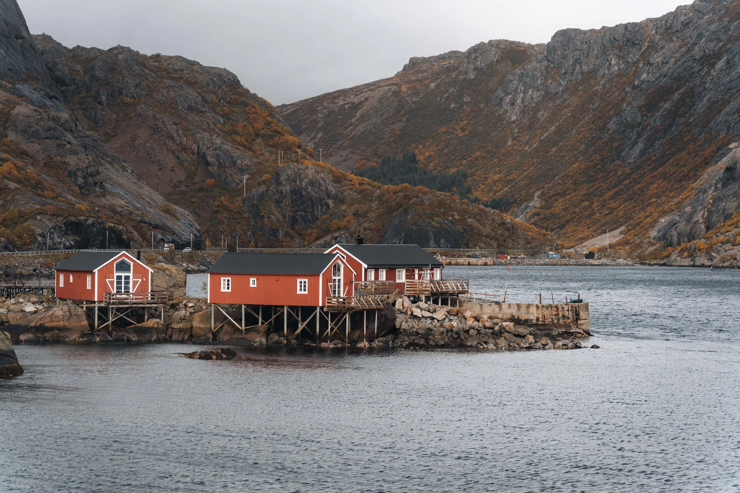 Rote Rorbuer auf Stelzen auf einem Felsen im Hafen von Nusfjord, dahinter die Berge
