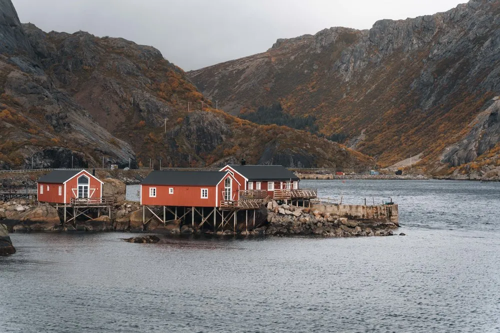 Rote Rorbuer auf Stelzen auf einem Felsen im Hafen von Nusfjord, dahinter die Berge