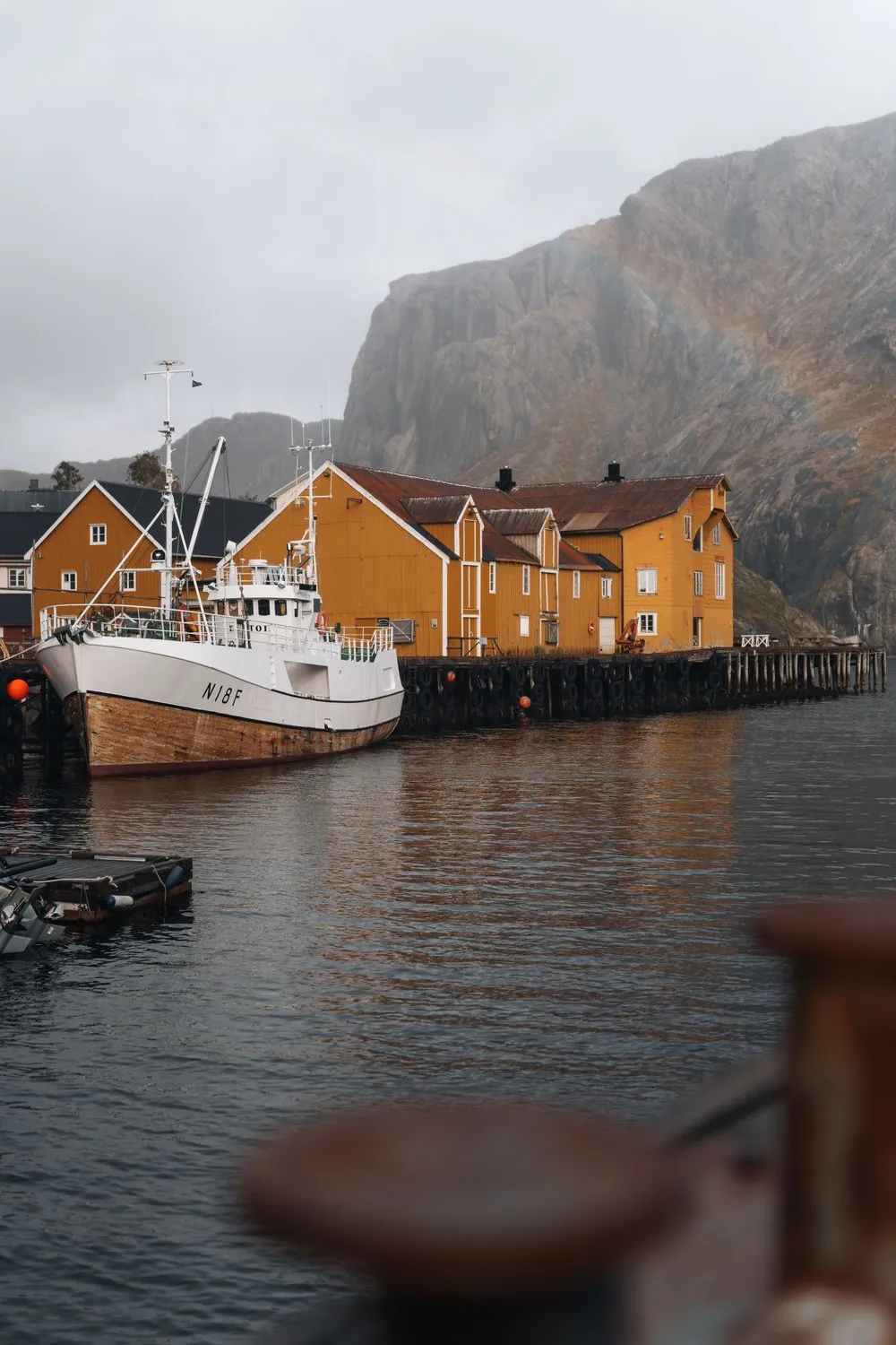 Ein Fischkutter vor den gelben Häusern am Kai von Nusfjord, darüber ein schwacher Regenbogen