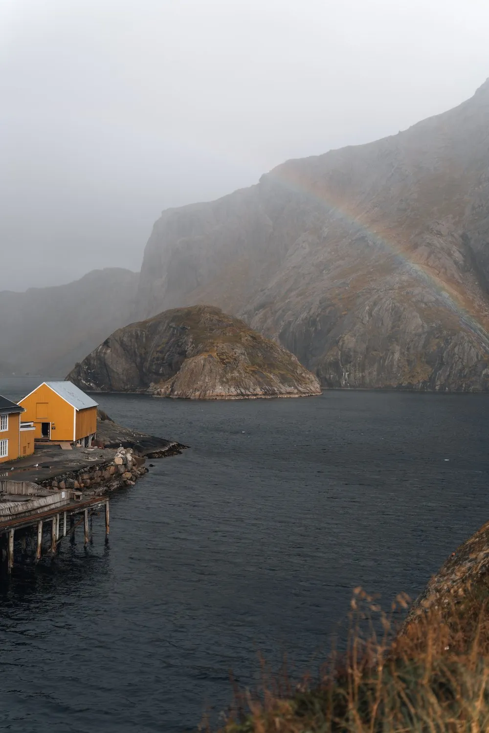 Ein Regenbogen über der Felswand bei Nusfjord, unten ein gelbes Haus am Wasser