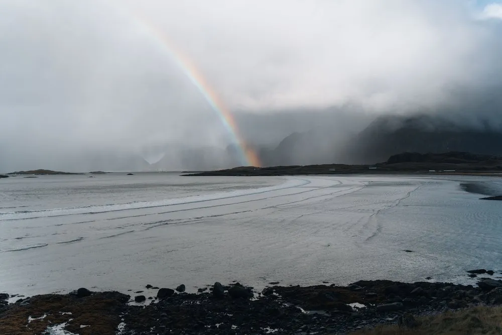 Der weite Sandstrand von Fredvang, darüber ein Regenbogen und Regenschauer über den Bergen