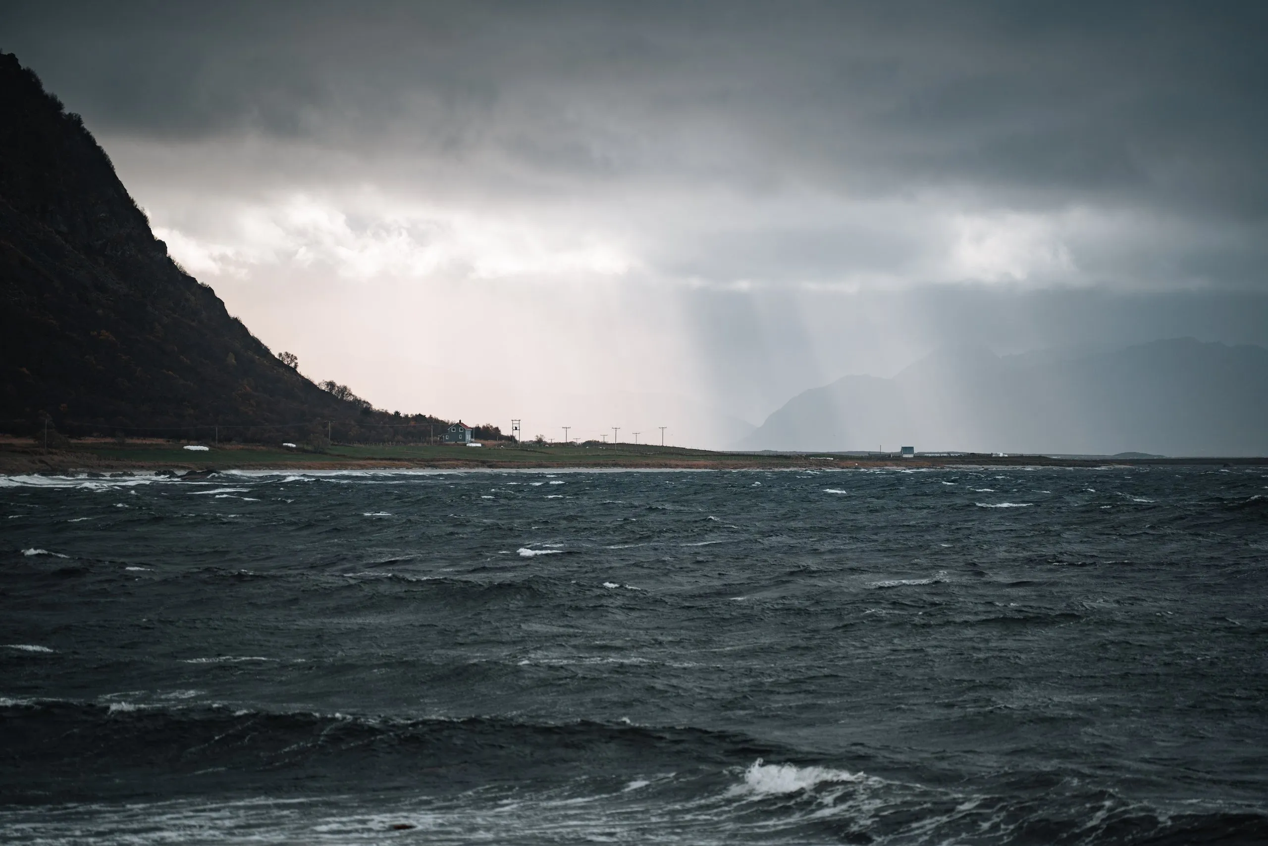 Regenschauer hängen als helle Schleier über dem aufgewühlten Meer bei Nøss, links ein dunkler Berghang, am Ufer ein einzelnes Haus