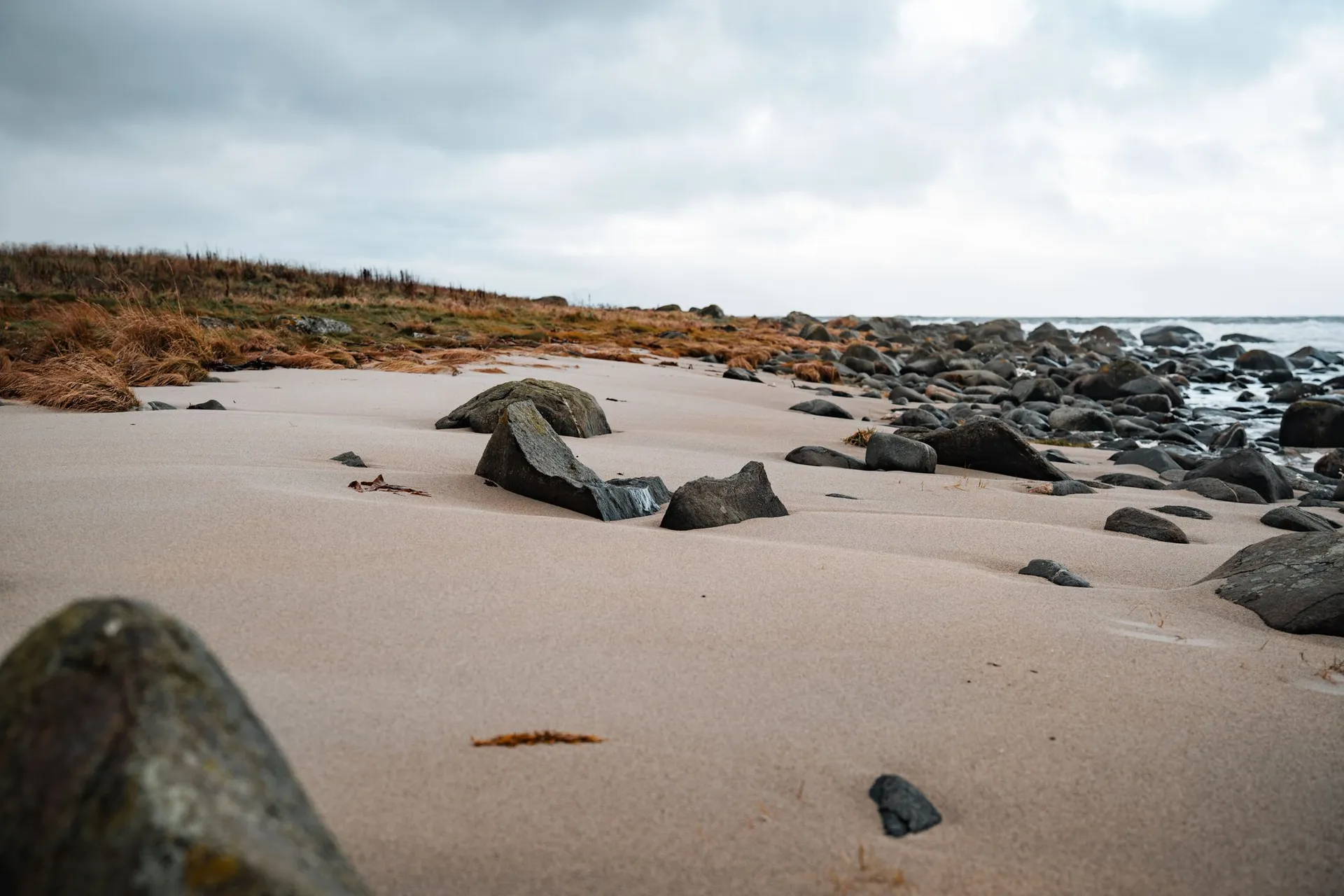 Dunkle, runde Steine im hellen Sand am Strand von Nøss, dahinter trockenes braunes Gras unter grauem Himmel