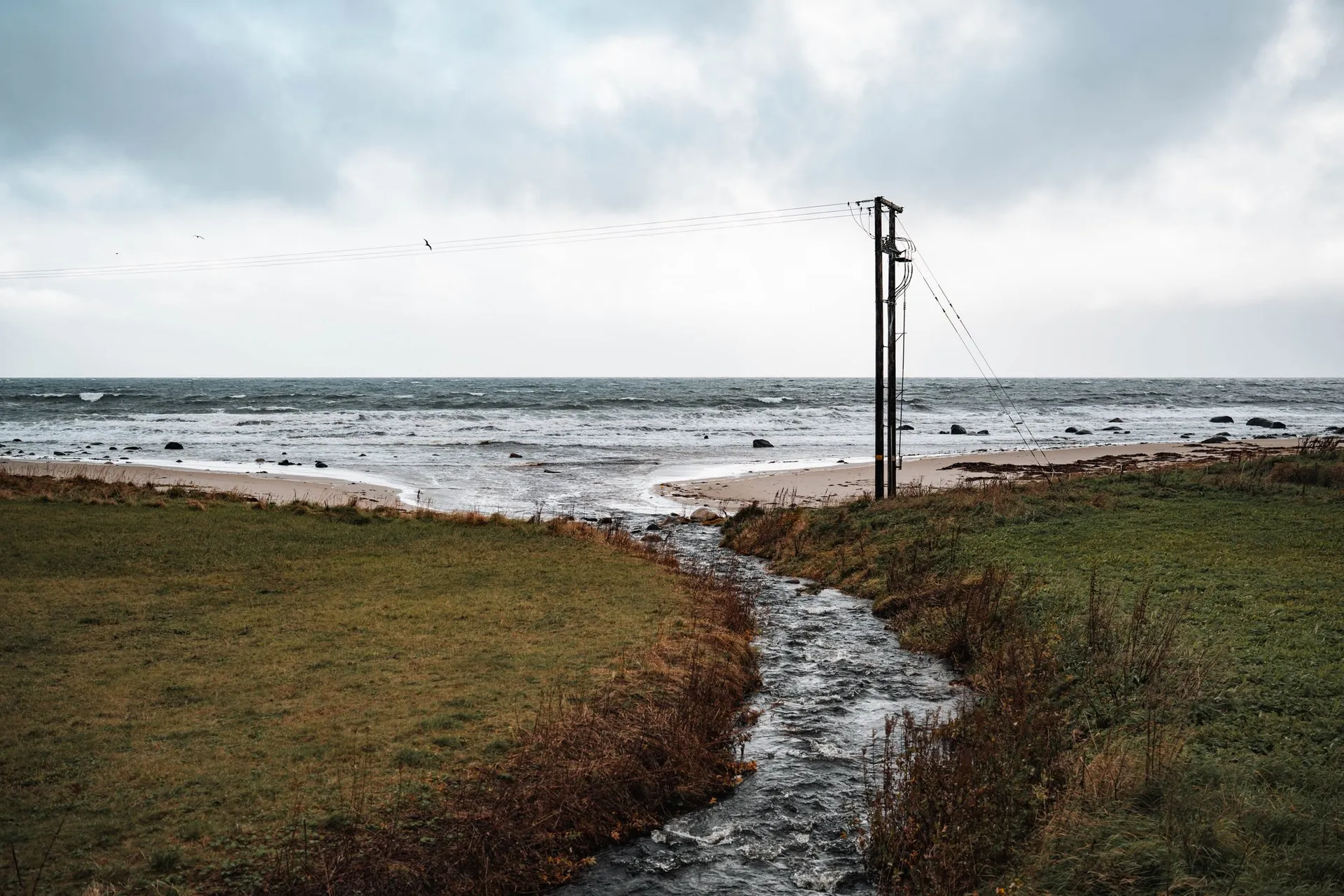 Ein Bach fliesst zwischen nassen Wiesen zum Strand von Nøss, daneben ein hölzerner Strommast, dahinter das aufgewühlte Meer