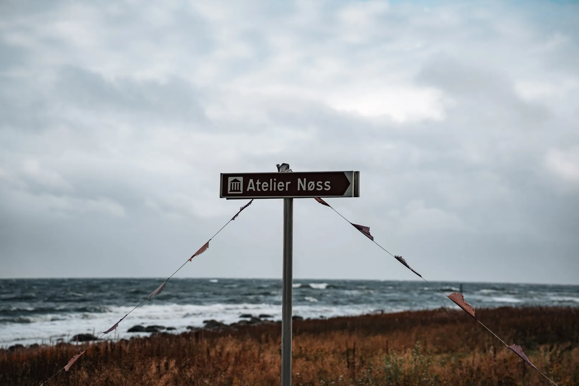 Ein braunes Wegweiser-Schild zum Atelier Nøss mit Wimpeln an zwei Schnüren, dahinter das Meer und braunes Gras
