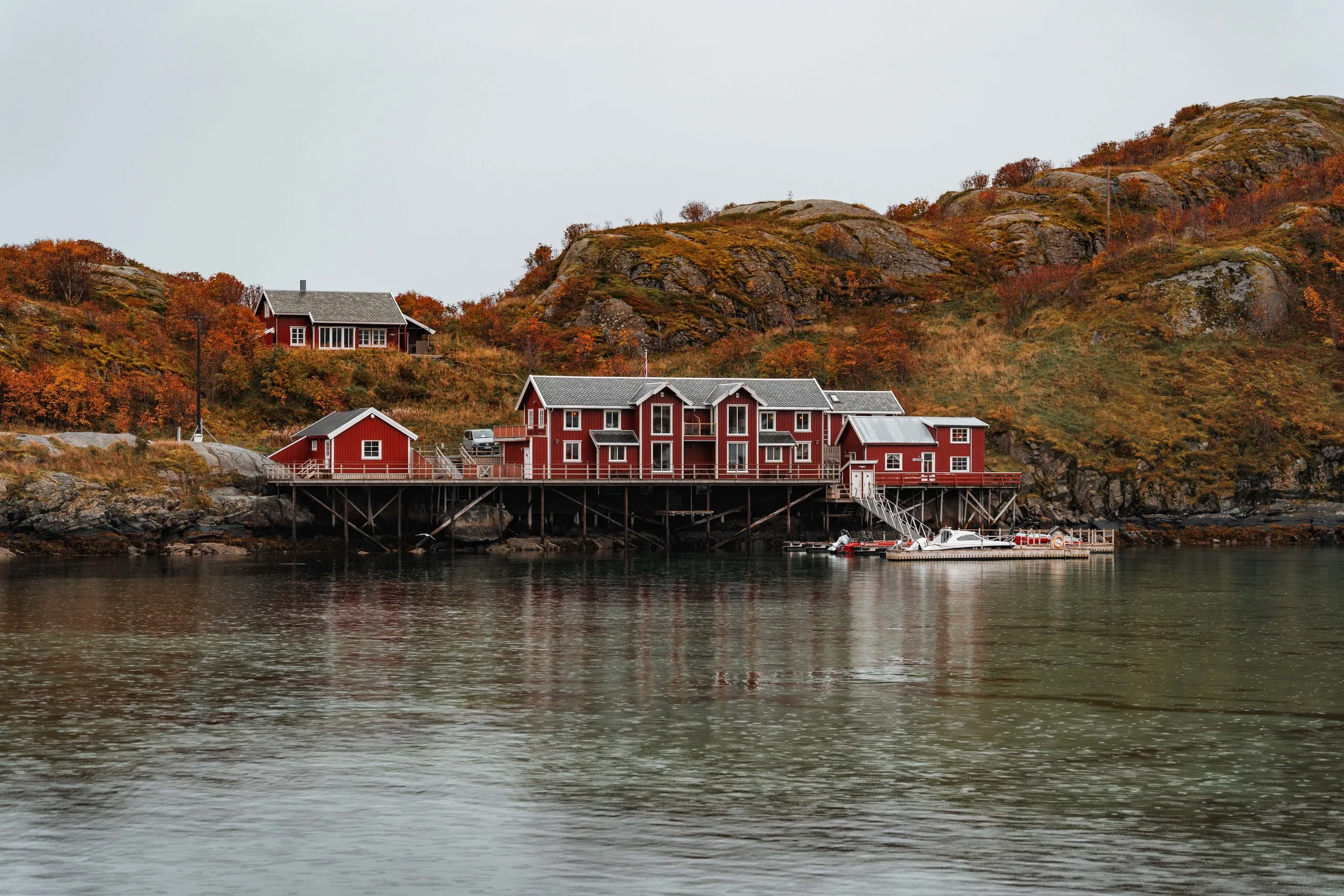 Rote Rorbuer auf Stelzen über dem ruhigen Wasser bei Sakrisøy, dahinter herbstlich orange Hügel