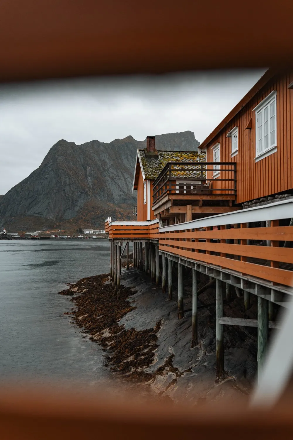 Durch eine Lücke zwischen Holzbalken auf die ockerfarbenen Rorbuer von Sakrisøy auf ihren Stelzen am Wasser