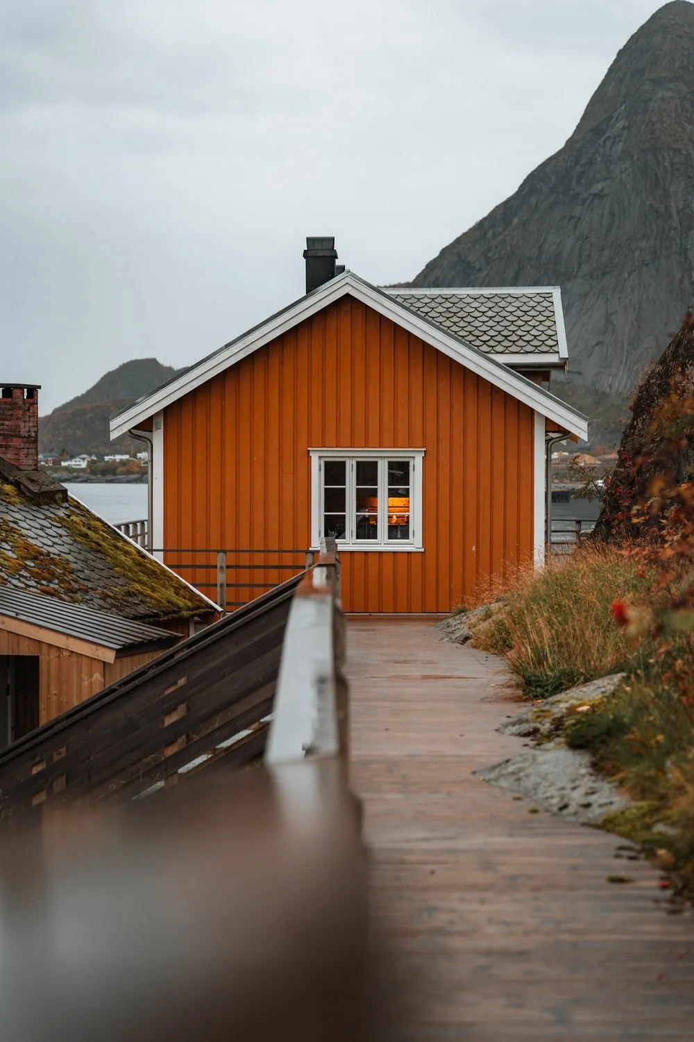Ein nasser Holzsteg führt zu einem ockerfarbenen Haus in Sakrisøy, dahinter ein spitzer Berg