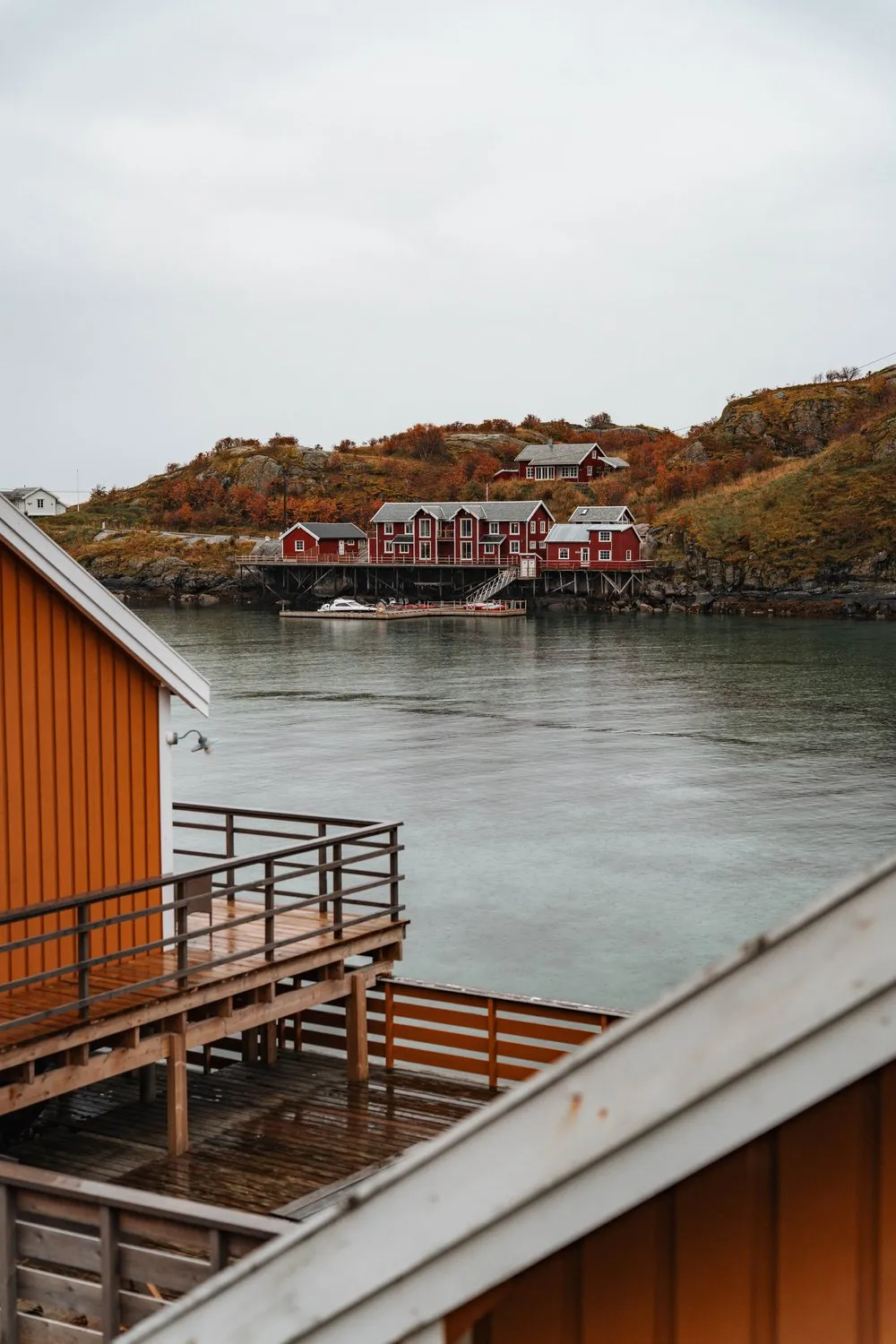 Von einem ockerfarbenen Rorbu in Sakrisøy über die Bucht auf rote Rorbuer vor herbstlichen Hügeln