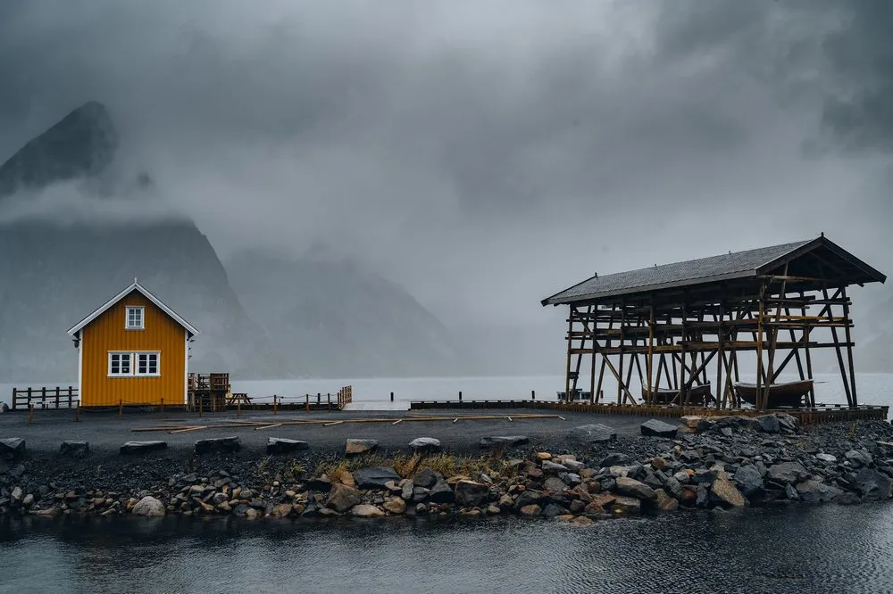Ein gelbes Fischerhaus und ein hölzernes Gestell mit Dach am Wasser in Sakrisøy, dahinter ein Berg im Nebel