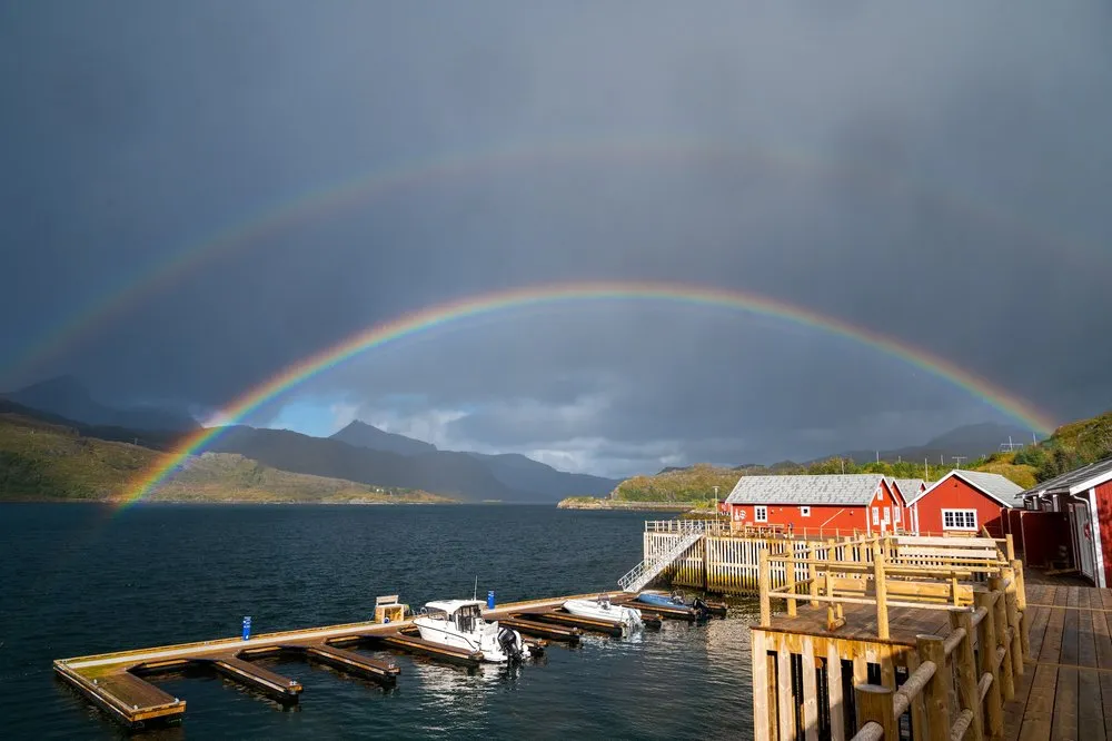 Ein doppelter Regenbogen über einem Bootssteg und roten Fischerhütten am Fjord