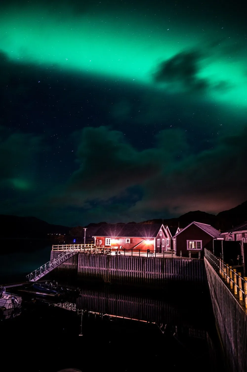 Ein grünes Nordlicht über beleuchteten roten Hütten und einem Steg am Wasser