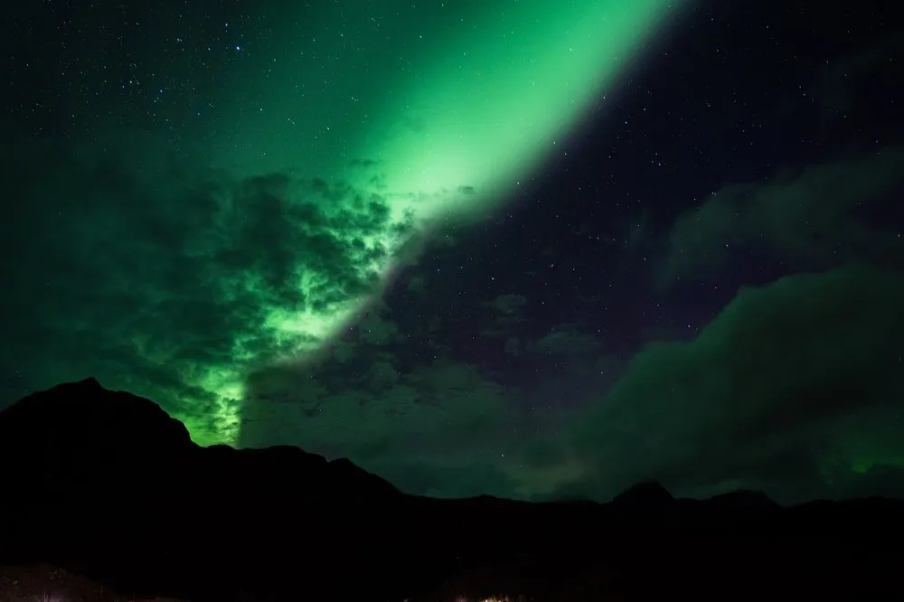 Ein grünes Nordlichtband steigt hinter den Umrissen dunkler Berge und Wolken auf