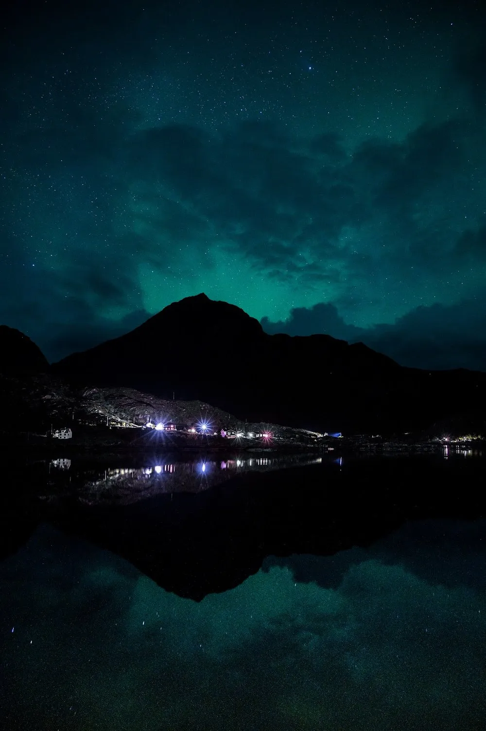 Ein Berg und die Lichter am Ufer spiegeln sich im stillen Wasser, darüber ein schwaches grünes Leuchten