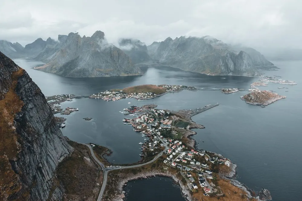 Blick aus grosser Höhe auf eine Landzunge mit kleinen Häusern, einer Strasse und einem Fjord, dahinter Berge in tiefen Wolken