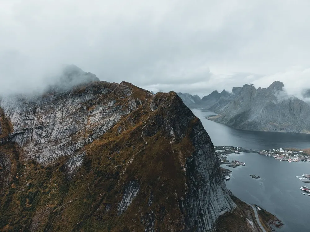 Steile, bewachsene Felsflanke ragt in die Wolken, rechts liegt ein Fjord mit einem Dorf am Ufer