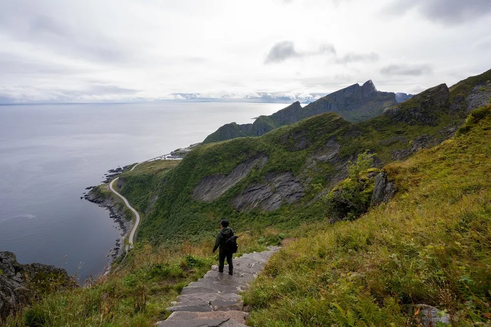 Eine Person mit Rucksack steht auf einer Steintreppe am grünen Hang, links das Meer und eine Strasse entlang der Küste