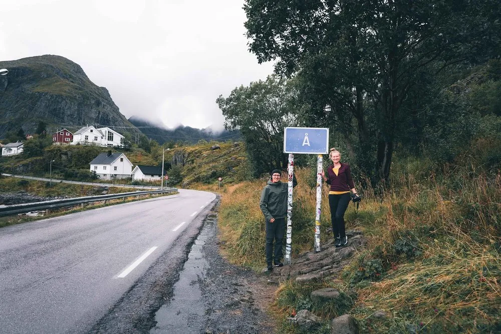 Zwei Personen stehen neben dem Ortsschild Å, dessen Pfosten voller Aufkleber ist, links eine Strasse und Häuser am Hang