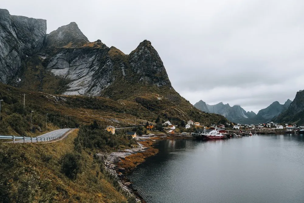 Eine Strasse führt am Hang entlang zu einem Dorf mit Hafen am Wasser, dahinter Felsgipfel unter grauem Himmel