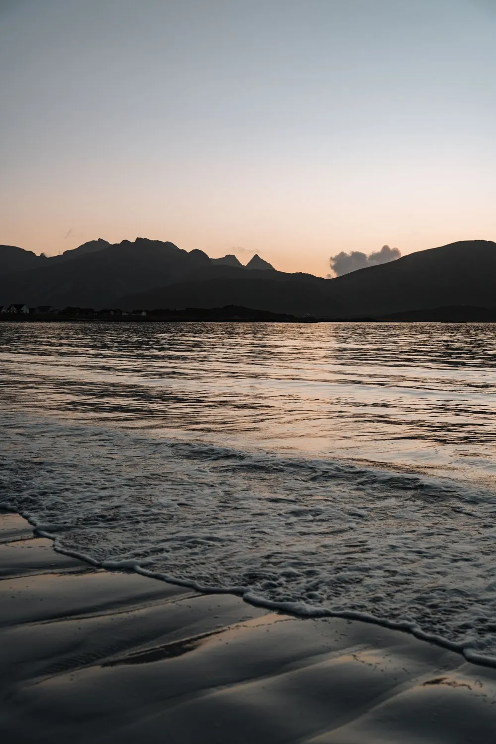 Eine flache Welle läuft über den nassen Sand am Rambergstranda, dahinter die Berge im letzten Licht