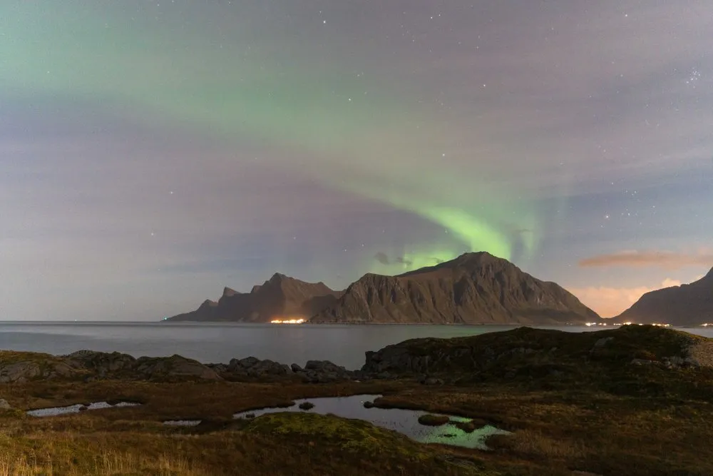 Ein grüner Nordlichtbogen über einem Berg am Meer, das Licht spiegelt sich in einem Tümpel im Vordergrund