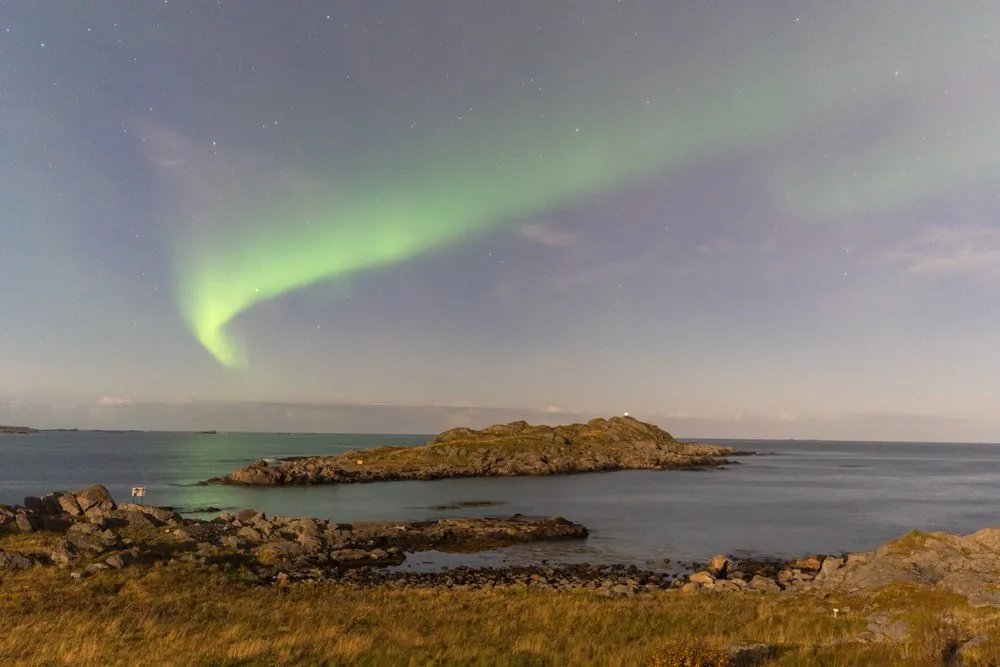Ein grünes Nordlichtband über einer flachen Insel im Meer, vorne Felsen und Gras
