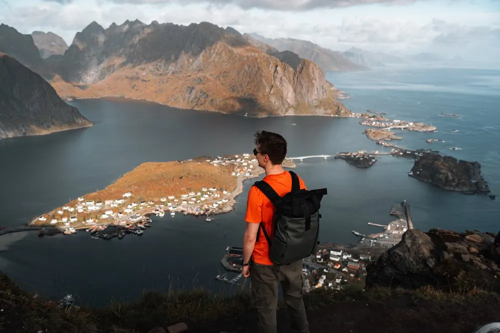 Nicolas im orangen Shirt mit Rucksack auf dem Reinebringen, unter ihm Reine und die Inseln im Fjord