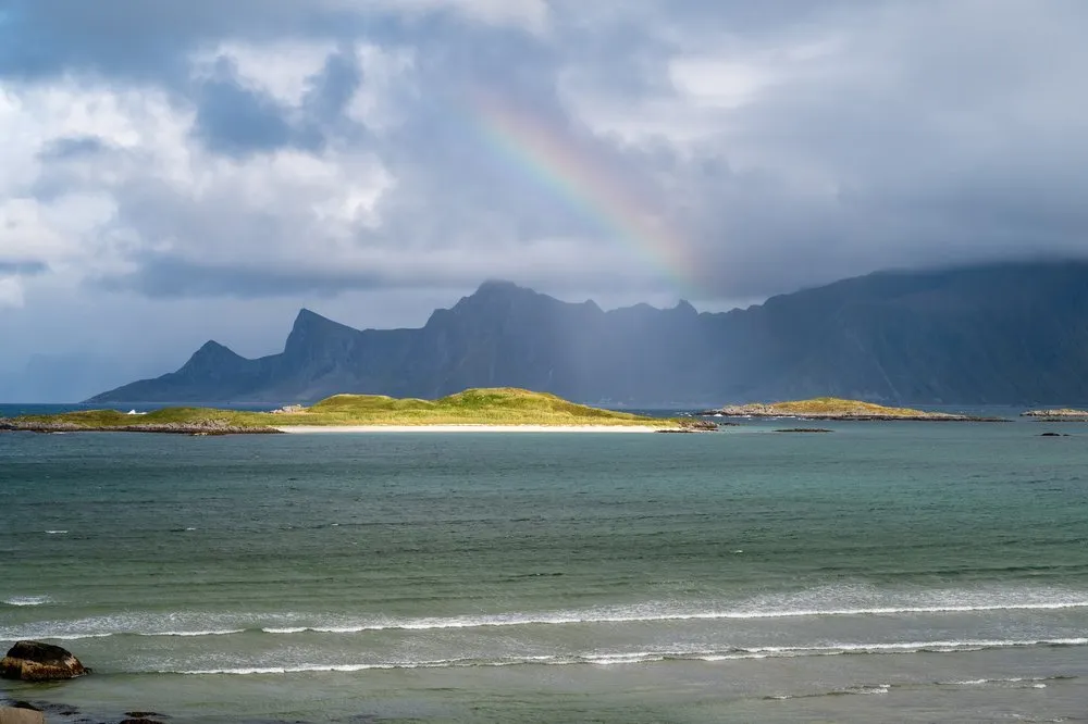Ein Regenbogen über den Bergen, davor türkisfarbenes Wasser und eine grasbewachsene Insel mit hellem Sandstreifen bei Fredvang