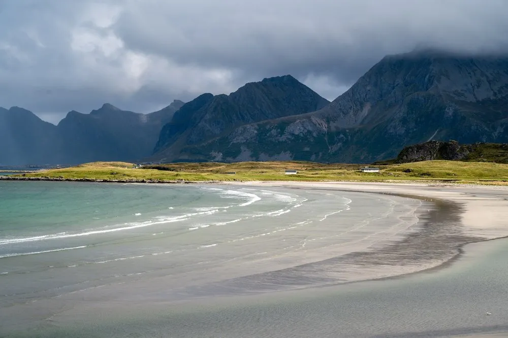 Ein geschwungener heller Sandstrand mit flachem Wasser, dahinter dunkle Berge unter Wolken
