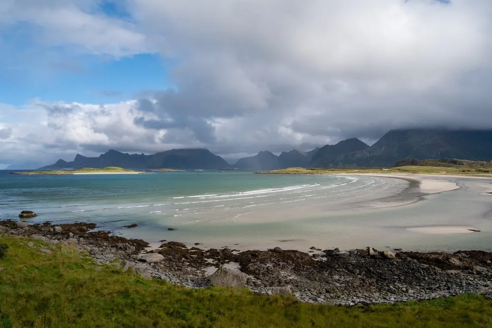 Eine Bucht mit hellem Sand und türkisfarbenem Wasser, im Vordergrund Steine und Gras, hinten Berge und blauer Himmel