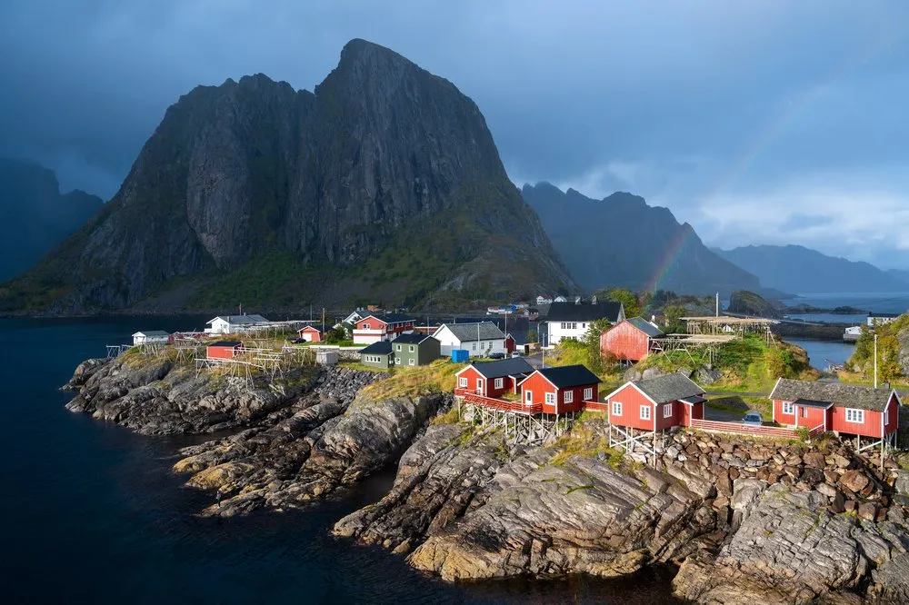 Rote Fischerhäuschen von Hamnøy auf Felsen am Wasser vor einem steilen Berg, dahinter ein Regenbogen unter dunklem Himmel
