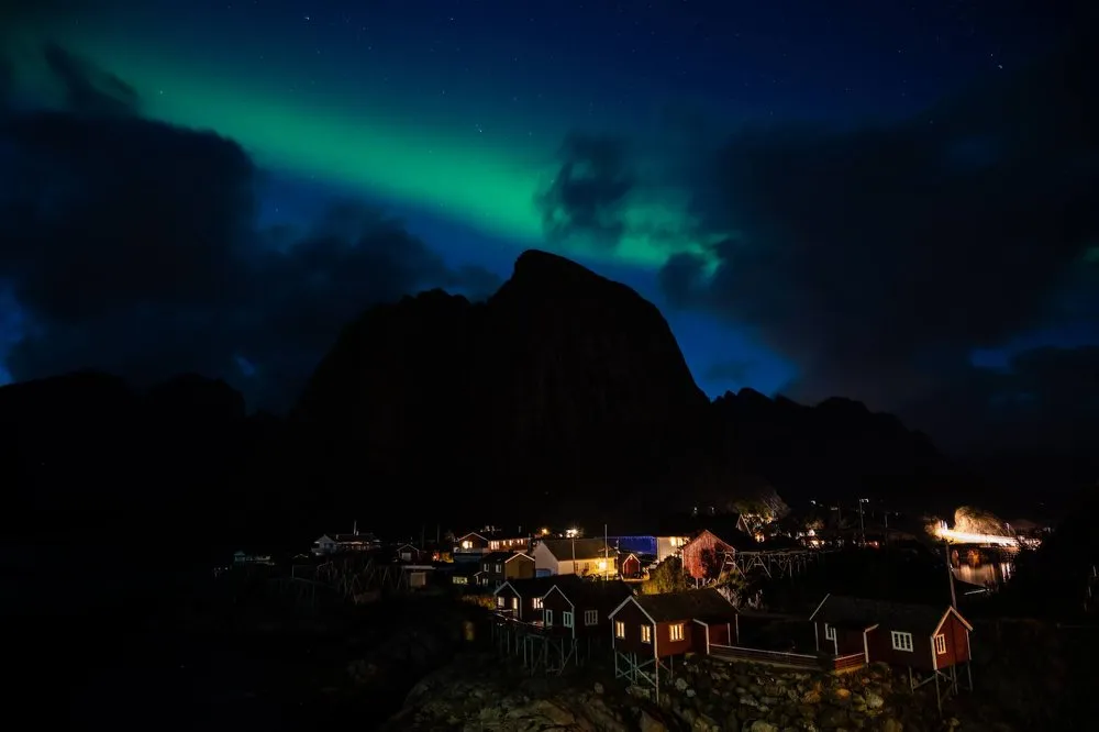 Grünes Nordlicht über der dunklen Silhouette des Bergs, unten beleuchtete rote Häuschen von Hamnøy