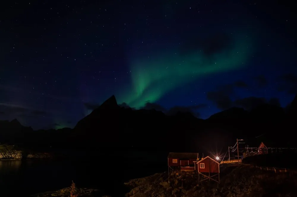 Nordlicht hinter einer spitzen Bergsilhouette, unten zwei rote Hütten am Wasser und ein heller Lichtpunkt
