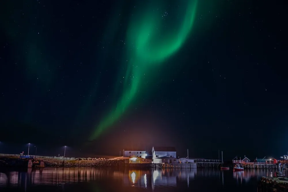 Ein grünes Nordlichtband über dem Hafen, davor weisse Gebäude und Strassenlampen, die sich im Wasser spiegeln