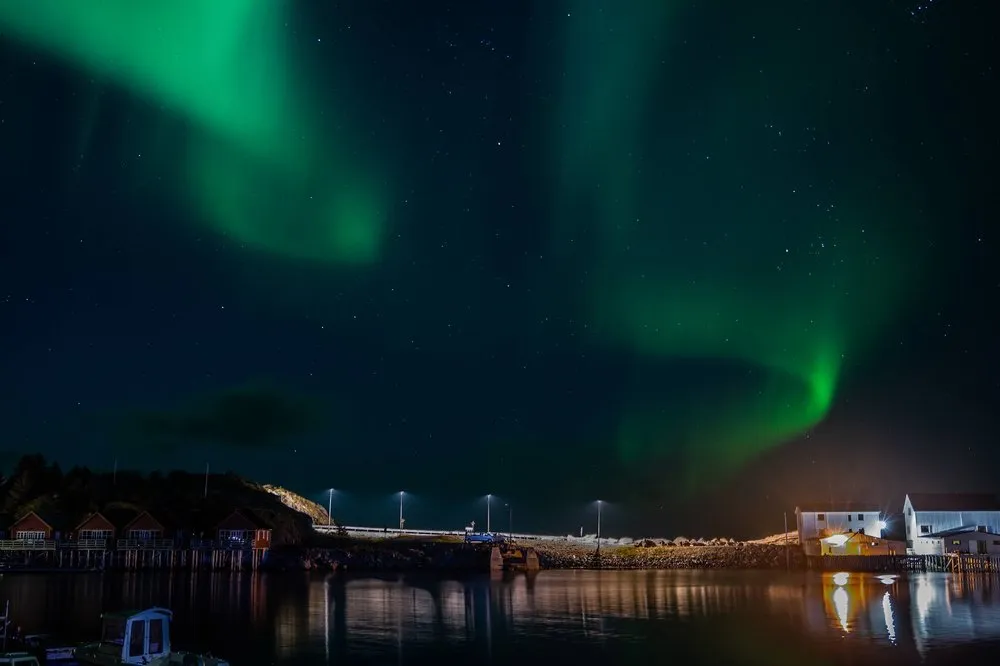 Grünes Nordlicht über dem Hafen, links Hütten unter einem Hügel, rechts weisse Gebäude, alles spiegelt sich im Wasser