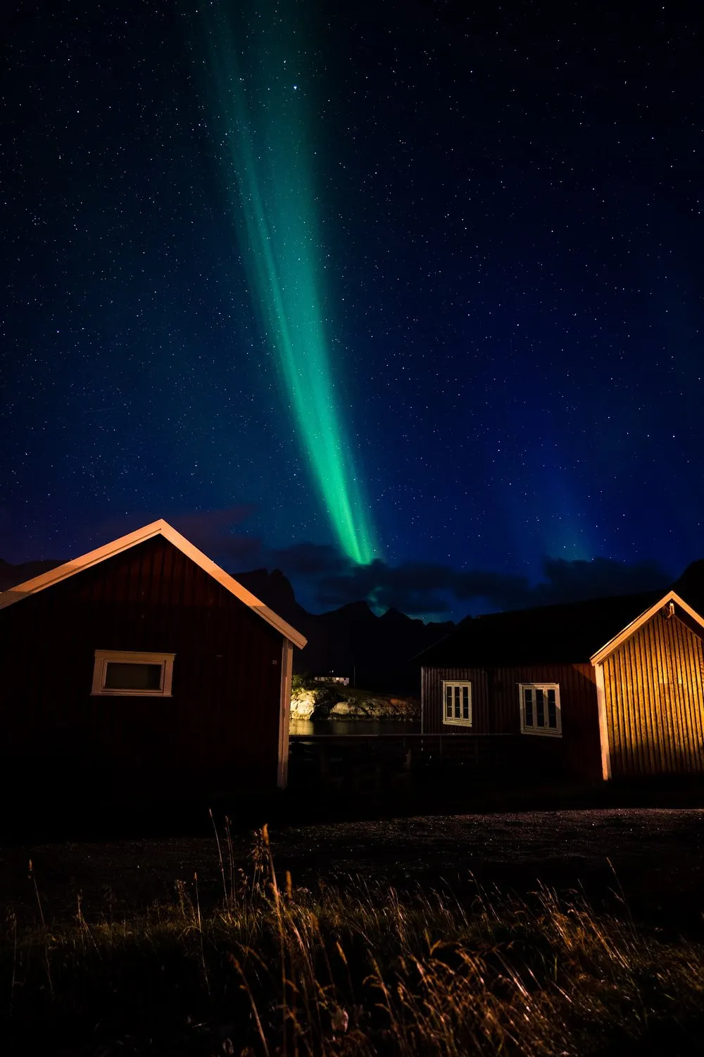 Ein schmaler grüner Lichtstrahl des Nordlichts steht zwischen zwei roten Hütten am Sternenhimmel