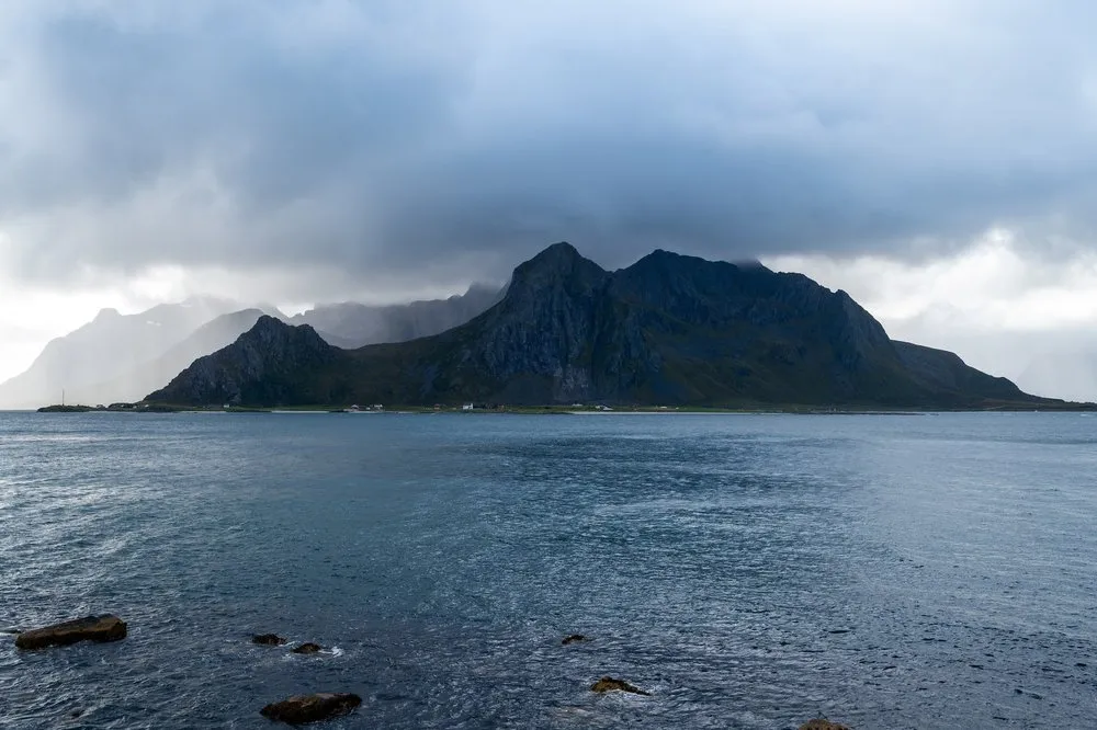 Eine dunkle Berginsel im Meer unter schweren Regenwolken, im Hintergrund ein heller Wolkenstreifen