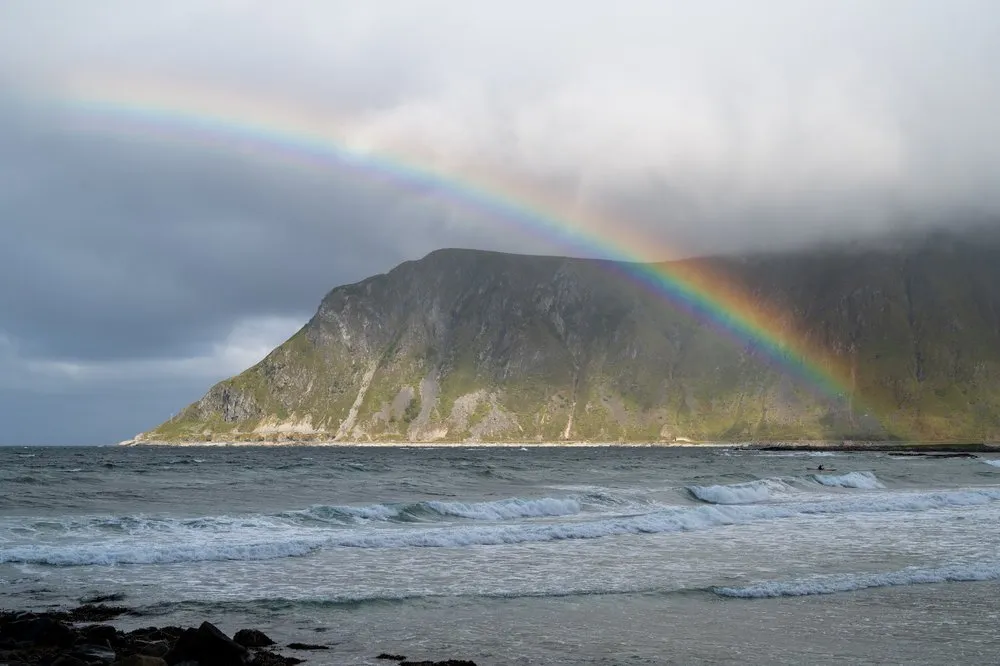 Ein Regenbogen spannt sich über einen grünen Berghang am Skagsanden Beach, davor rollen Wellen an den Strand