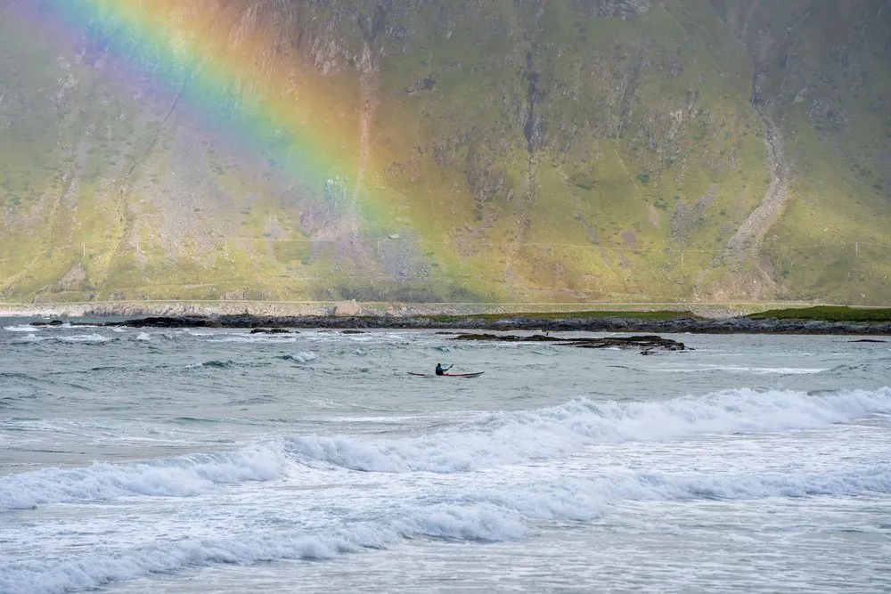 Ein Kajakfahrer in der Brandung am Skagsanden Beach, dahinter ein Berghang mit dem Ende eines Regenbogens