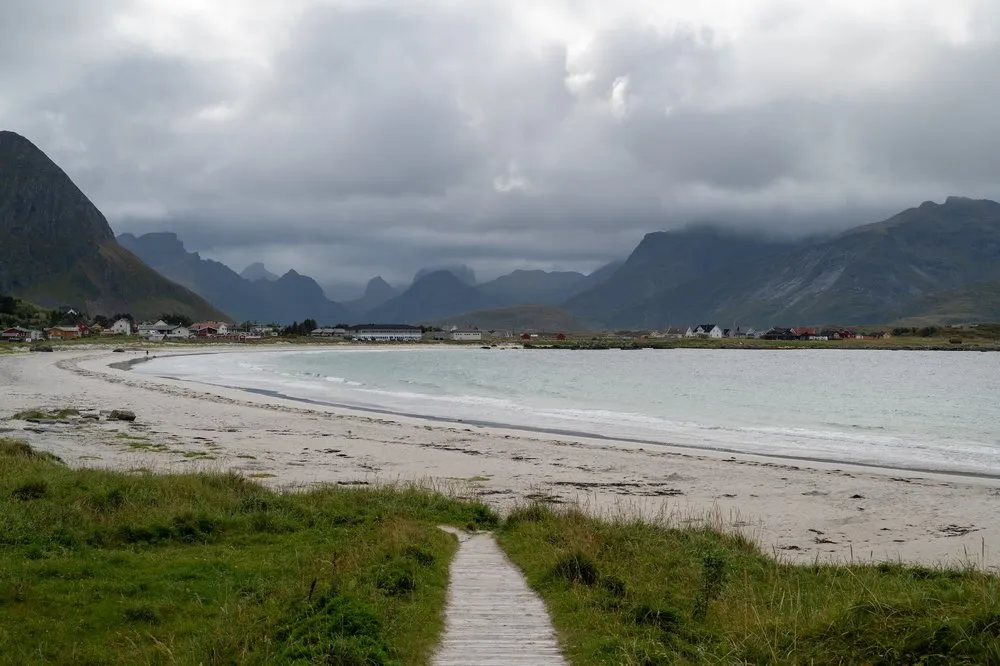 Ein Holzsteg führt durch Gras zu einem hellen Sandstrand am Rambergstranda, dahinter Häuser und Berge in Wolken
