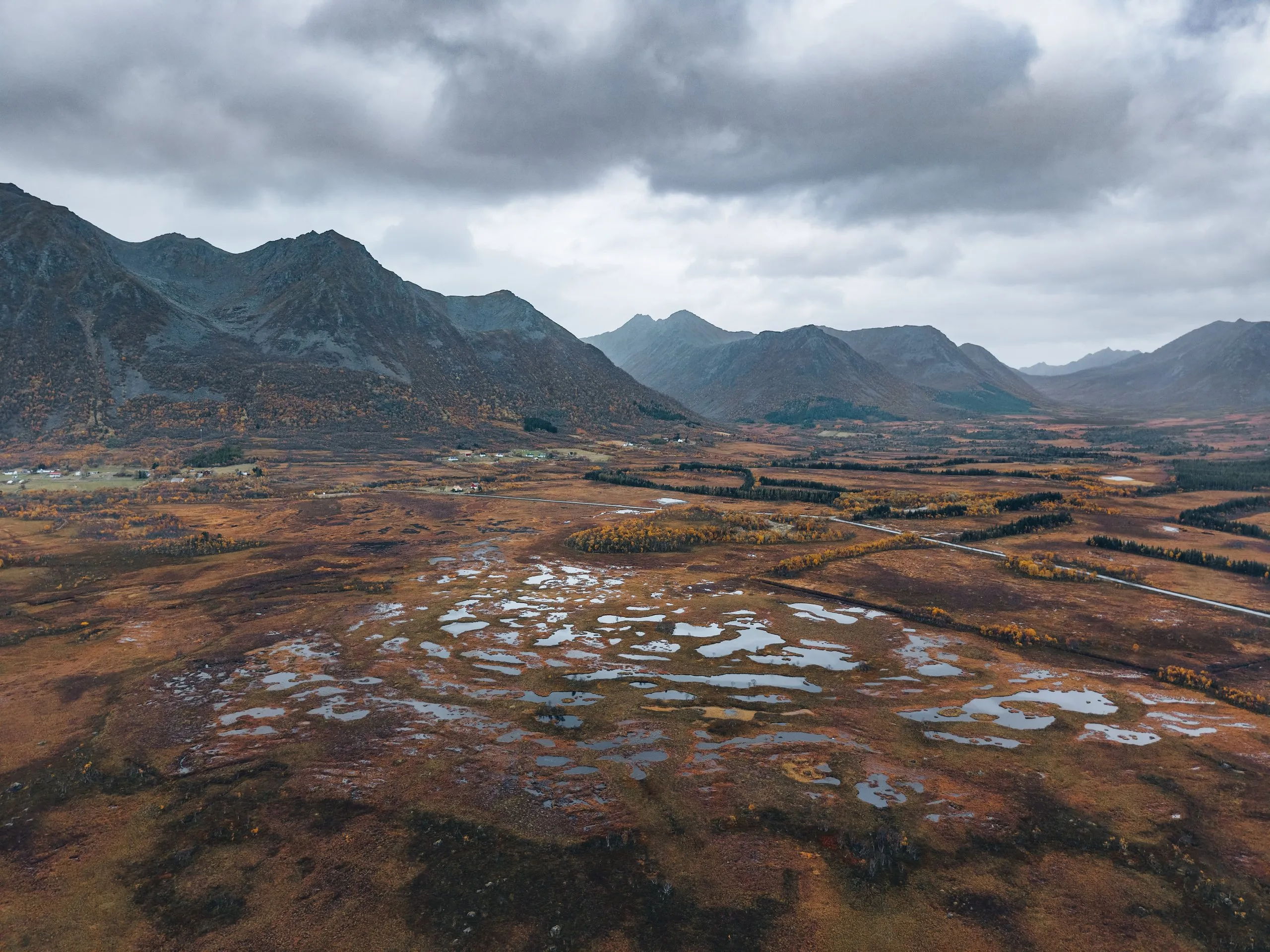 Ein rotbraunes Moor mit vielen kleinen Tümpeln aus der Luft, dahinter eine Bergkette unter grauen Wolken, unterwegs nach Andøya