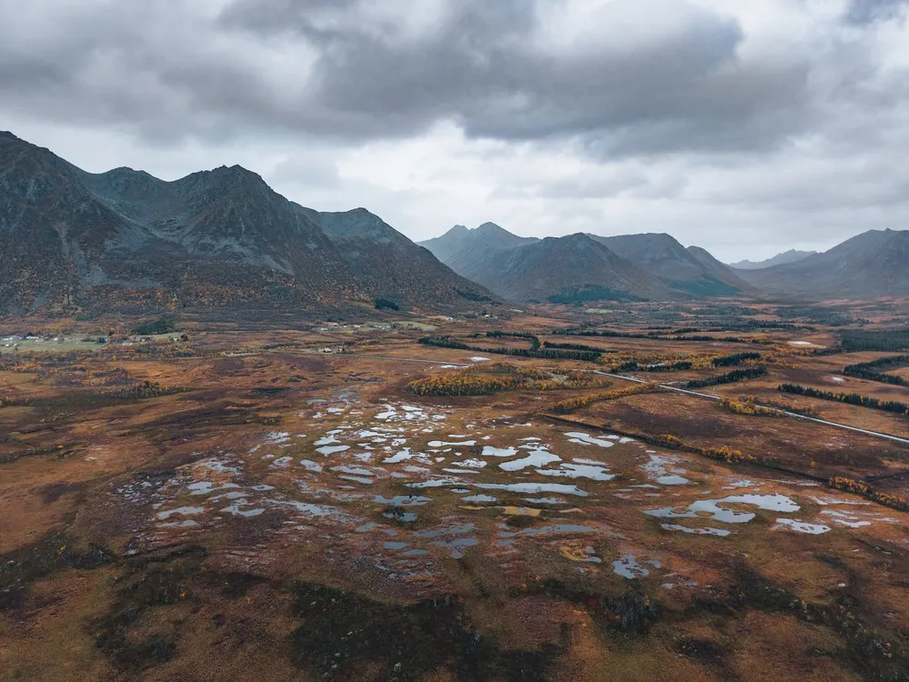 Ein rotbraunes Moor mit vielen kleinen Tümpeln aus der Luft, dahinter eine Bergkette unter grauen Wolken, unterwegs nach Andøya