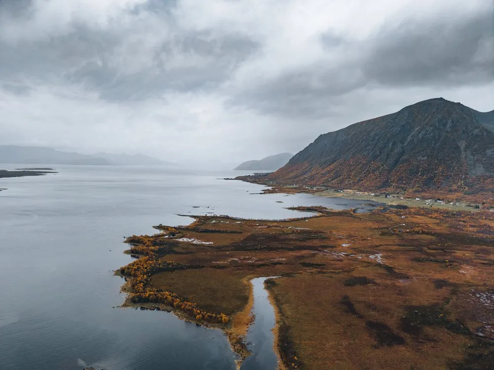 Eine flache Landzunge mit rotbraunem Moor und einem Bach, daneben ein dunkler Berg am Fjord, aus der Luft unterwegs nach Andøya